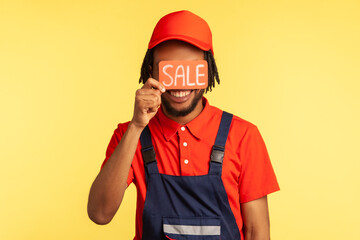 Unknown male worker wearing blue overalls and red T-shirt, standing covering eyes with card with...