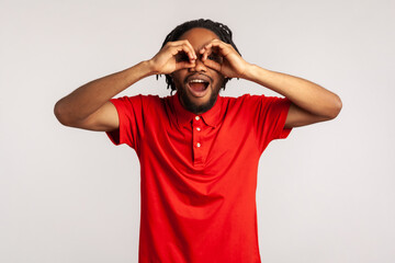 Amazed young adult man with dreadlocks wearing red casual style T-shirt, looking through fingers shaped like binoculars and expressing excitement. Indoor studio shot isolated on gray background. - obrazy, fototapety, plakaty