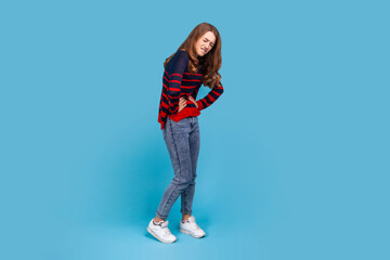 Full length portrait of woman wearing striped casual style sweater, frowning face from pain, suffering from painful period, gastritis or constipation. Indoor studio shot isolated on blue background.