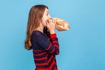 Side view of nervous stressed woman wearing striped casual style sweater, hardly breathing using paper bag, trying to calm down, to improve well-being. Indoor studio shot isolated on blue background. - obrazy, fototapety, plakaty