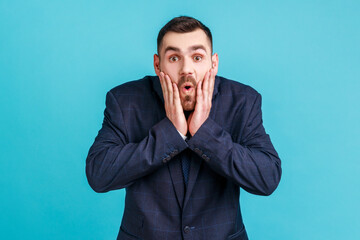Photo of amazed bearded young adult man in official style suit looks at camera with open mouth, raising palms, being impressed of shocking news. Indoor studio shot isolated on blue background.
