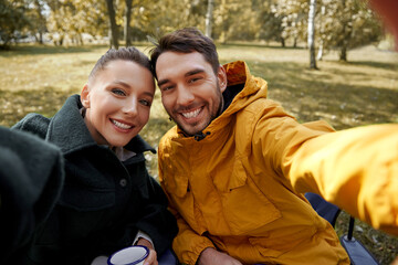 camping, tourism and travel concept - happy couple drinking tea and taking selfie at tent camp