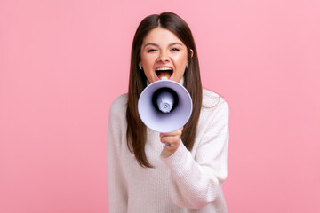 Portrait of excited female screaming in megaphone, announcing important information with positive, wearing white casual style sweater. Indoor studio shot isolated on pink background.