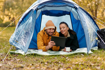 camping, tourism and travel concept - happy couple with tablet pc computer lying inside tent and drinking tea at campsite