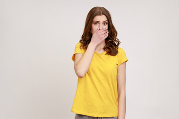 I won't say anyone! Frightened teenager girl in yellow T-shirt covering her mouth with arm, looking at camera amazed, shocked by breathtaking secret. Indoor studio shot isolated on gray background.