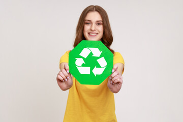 Smiling positive teenager girl with brown hair in casual style yellow t-shirt holding recycling green symbol, sorting rubbish, saving ecology. Indoor studio shot isolated on gray background.