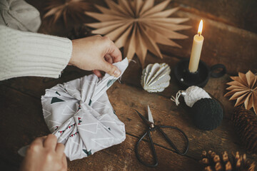 Furoshiki Christmas gift. Hands wrapping christmas gift in modern fabric on rustic wooden table with scissors, craft paper star, candle. Atmospheric moody time, nordic style. Zero waste holiday - obrazy, fototapety, plakaty