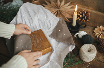 Hands wrapping christmas gift box in brown fabric on rustic wooden table with scissors, craft paper star, candle. Zero waste and eco friendly presents. Atmospheric moody time, nordic style. - obrazy, fototapety, plakaty