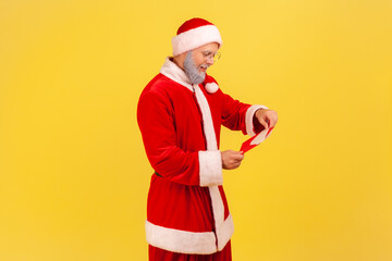 Fototapeta na wymiar Side view of elderly man with gray beard in santa claus costume standing open red envelope with congratulations with winter holidays , reading letter. Indoor studio shot isolated on yellow background.