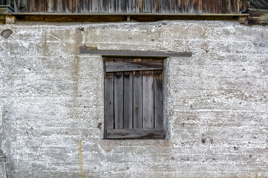There is a boarded-up wooden window on the old weathered concrete wall. An abandoned village barn.