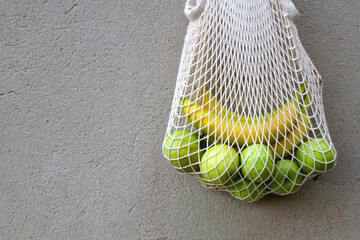 Crochet bag filled with green apples and bananas, hanging on a concrete wall.