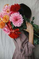 Beautiful woman holding colorful dahlias flowers in rustic room. Florist in linen dress hiding behind beautiful autumn bouquet. Atmospheric aesthetic image. Autumn season in countryside