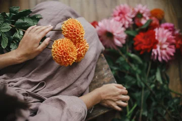 Schilderijen op glas Woman holding orange dahlia flower and sitting on wooden rustic bench, view above. Atmospheric moody image. Florist in linen dress arranging autumn flowers bouquet in countryside. Slow life © sonyachny