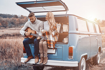 Handsome young man playing guitar for his girlfriend while both sitting in the car trunk outdoors