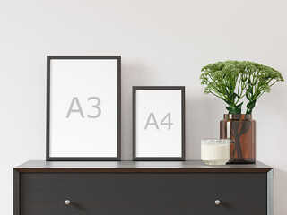 Two vertical empty A3 and A4 frames on white wall with black dresser, green plant in brown glass vase - close up image.  - obrazy, fototapety, plakaty