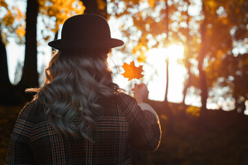 Autumn, fallen leaves, hello october concept. Back view of young hipster woman in hat holding and watching at fallen orange maple leaf in front of sunset sun outdoors. Selective focus on girl in coat
