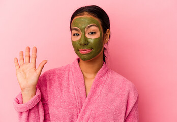 Young Venezuelan woman wearing a bathrobe and facial mask isolated on pink background smiling cheerful showing number five with fingers.