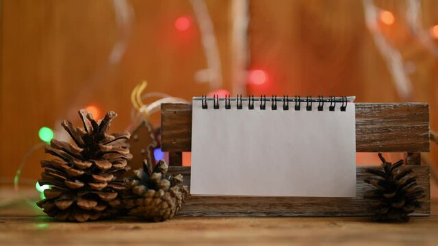 Notepad with blank white page in Christmas decorations and colorful garland lights. A horizontal blank page to add your own label. Selective focus on notepad.