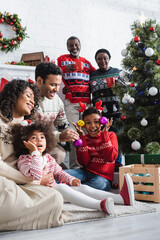 excited boy in reindeer horns headband holding decorative baubles near christmas tree and smiling african american family