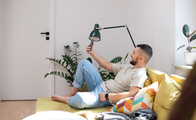 guy in casual clothing holding and using smartphone while lying on sofa in apartment.