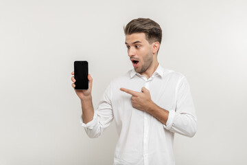 Young handsome happy man wearing white shirt, holding blank screen smartphone, pointing and watching at mobile, isolated over white background