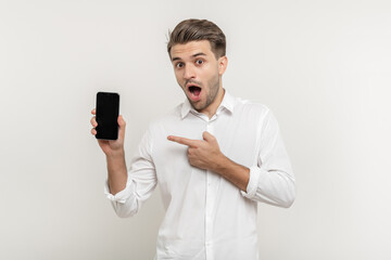 Portrait of surprised brunette man with beard in white shirt pointing at cellphone and screaming at camera, recommending gadget or mobile application. indoor studio shot isolated on white background