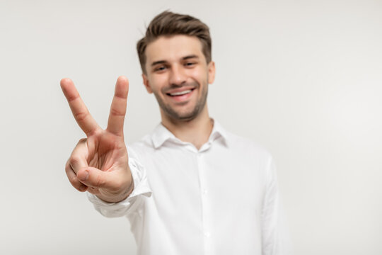 Young happy man with beard wearing white shirt standing over white background smiling and showing two fingers on camera hand doing victory sign. Number two. focus on hand