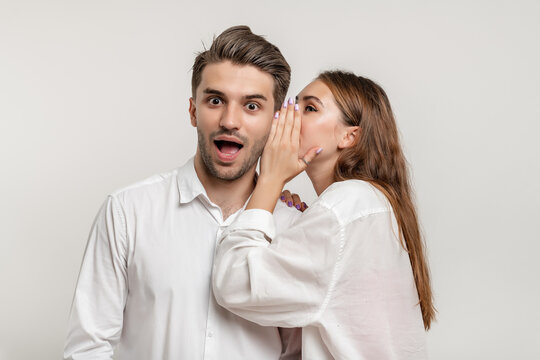 Image of beautiful young woman whispering secret or interesting gossip to shocked man in his ear isolated over white background