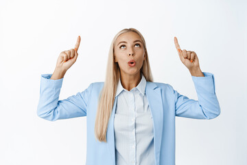 Portrait of blond saleswoman, pointing and looking up with amazed face expression, standing in blue suit over white background