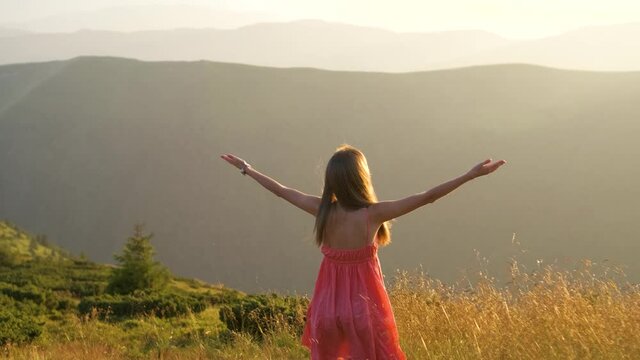 Back view of young happy woman traveler in red dress standing on grassy hillside on a windy evening in summer mountains with outstretched arms enjoying view of nature at sunset.