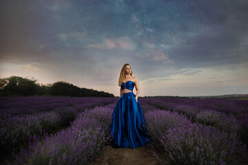 A blonde girl on a background of the starry sky in a lavender field dressed in a blue dress. Summer sunrise and a model posing in the lavender bushes.