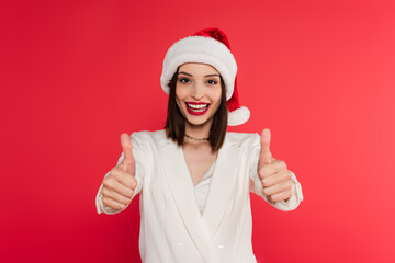 Positive woman with red lips and santa hat showing thumbs up isolated on red