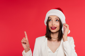 Smiling woman with red lips and santa hat talking on smartphone and pointing with finger isolated on red