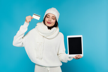 Smiling woman in cozy sweater and hat holding credit card and digital tablet with blank screen isolated on blue