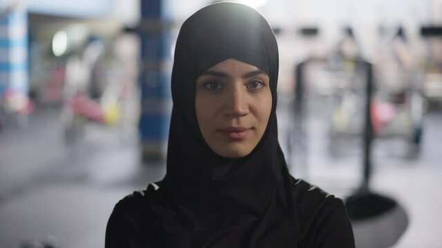 Portrait Of Gorgeous Young Beautiful Middle Eastern Woman In Hijab Turning To Camera Standing In Gym Light Indoors. Beautiful Slim Sportswoman Posing In Sport Club. Training And Fitness Concept