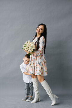 Photo portrait of a brunette girl holding a bouquet of white tulips in her hands, standing next to her four-year-old child, isolated against a gray background. Mother's Day