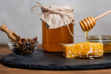 honeycomb, wooden dipper, bowl with anise seeds and blurred jar with fresh honey on slate board and grey background