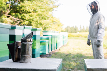 bee smoker on beehive near beekeeper in safety suit on apiary