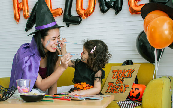 Asian beautiful woman or mother and little Caucasian sweet girl wearing witch costume with hats, playing and painting face with fun, smiling with happiness, celebrating Halloween party at home.