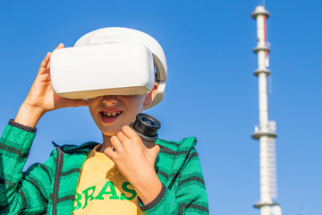 Obraz premium Child wearing virtual reality glasses and holding the camera in the left hand, in the background visible blue clear sky and mobile telephony relay