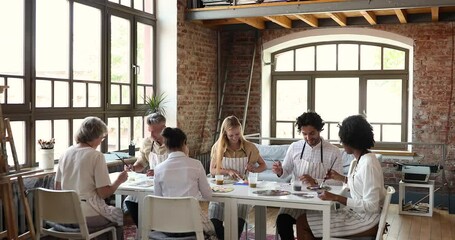 Inside loft workshop room group of six diverse people sit at table attend art therapy or artistic studio class, holding paintbrushes drawing on paper and talking. Hobby, education, creativity concept