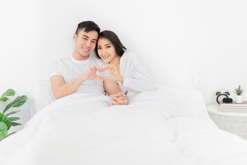Obraz na płótnie Canvas Asian lover show heart sign with hand, they feeling happy and smiling, they rest on bed, happiness honeymoon