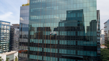 Two male workers with safety equipment at heights. Cleaning up a tall mirrored glass building by...