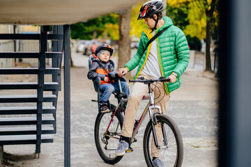 Cute little boy in child seat to ride a bicycle with his father in city street. Father walking with...