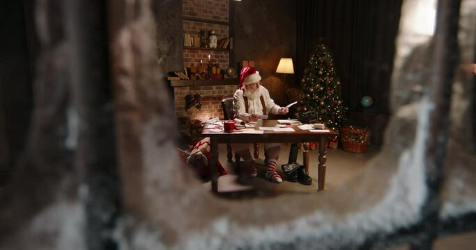 Busy Santa Claus reading kids letters, working at night, outside view through window. St Nicholas receiving children wishes. Late work in evening on Christmas Eve, Santa Claus Post office.