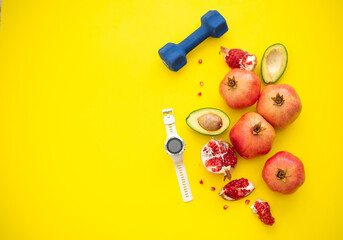 Lot of pomegranates, cut avocado, dumbbells and sport watch on yellow background. Fruits contain many vitamins. healthy fruits:  good for body. food for diet. top view. Place for text.