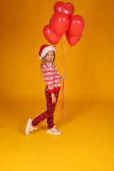 a little beautiful happy blonde girl in a New Year's jumper and plaid trousers with a Santa Claus hat holding red balloons on a yellow background