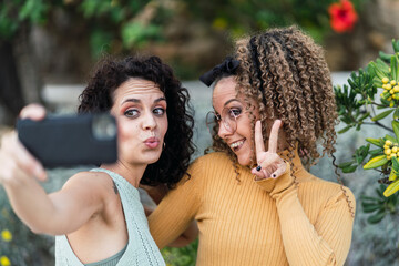 Dos amigas tomándose selfies en la calle junto a un jardin