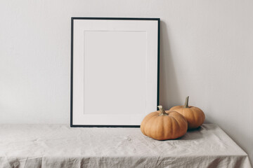 Autumn still life. Vertical black picture frame mockup. Pale orange pumpkins on linen table cloth. White wall background. Minimal rustic interior, neutral color. Halloween, Thanksgiving concept.