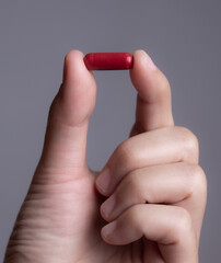 Male hand holding a medical red capsule pill between fingers against gray background